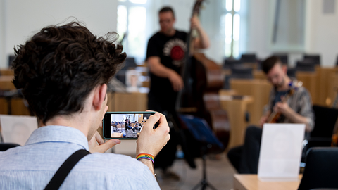 Zu sehen ist ein Besucher, der mit seinem Smartphone Musiker im Hintergrund aufnimmt.
