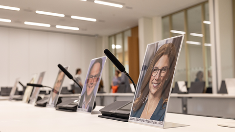 Zu sehen sind nebeneinder stehende Tische, auf denen Fotos von einigen Abgeordenten des Landtags stehen sowie dazwischenstehende Tischmikrophone.