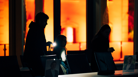 Zu sehen sind die Umrisse von drei Menschen, die durch einen Raum gehen. Im Hintergrund sieht man etwas Licht.