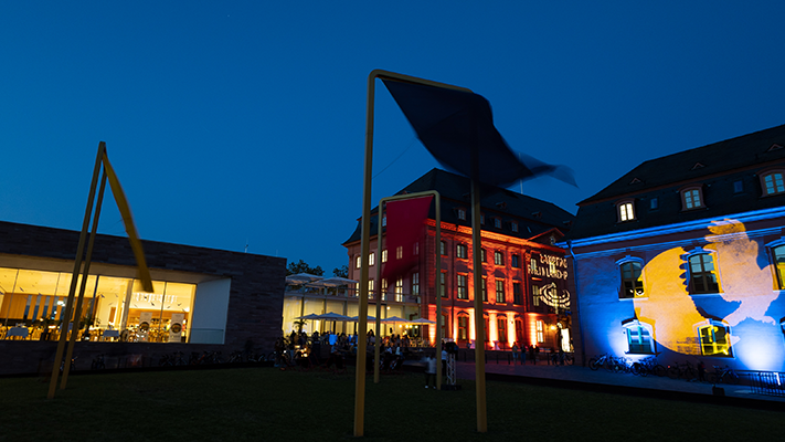 Zu sehen sind mehrere Gebäude bei Nacht, die angeleuchtet werden. Auf der rechten Seite sieht man das blau-gelbe Logo der Museumsnacht.
