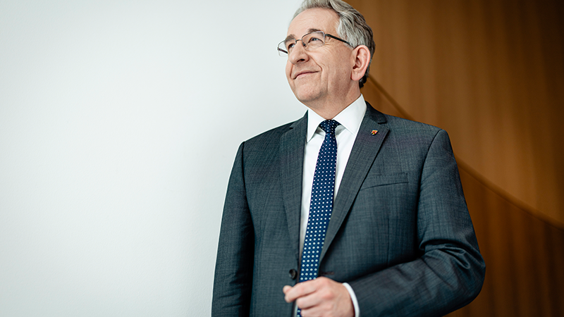 Zu sehen ist ein Mann im Profil stehend, mit leicht lächelndem Blick. 