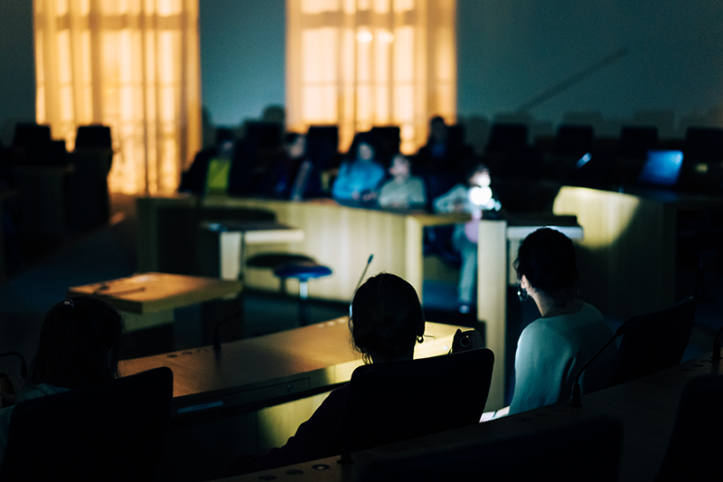 Zu sehen ist ein Teil des dunklen Plenarsaals. Man sieht die Umrisse einiger sitzenden Menschen.