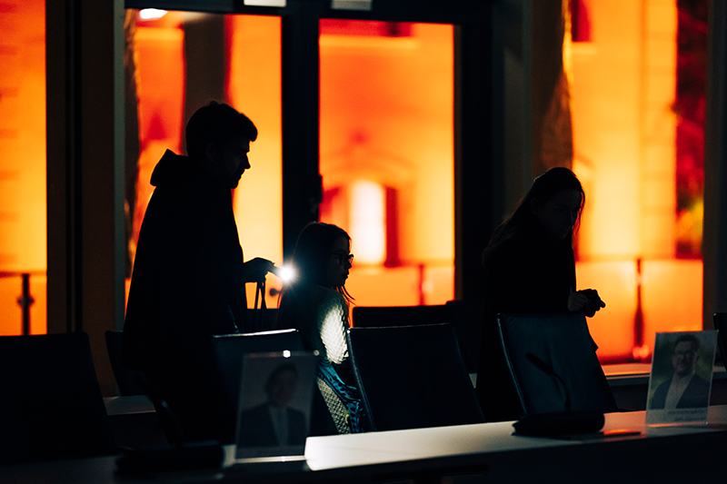 Zu sehen sind die Umrisse von drei Menschen, die durch einen Raum gehen. Im Hintergrund sieht man etwas Licht.