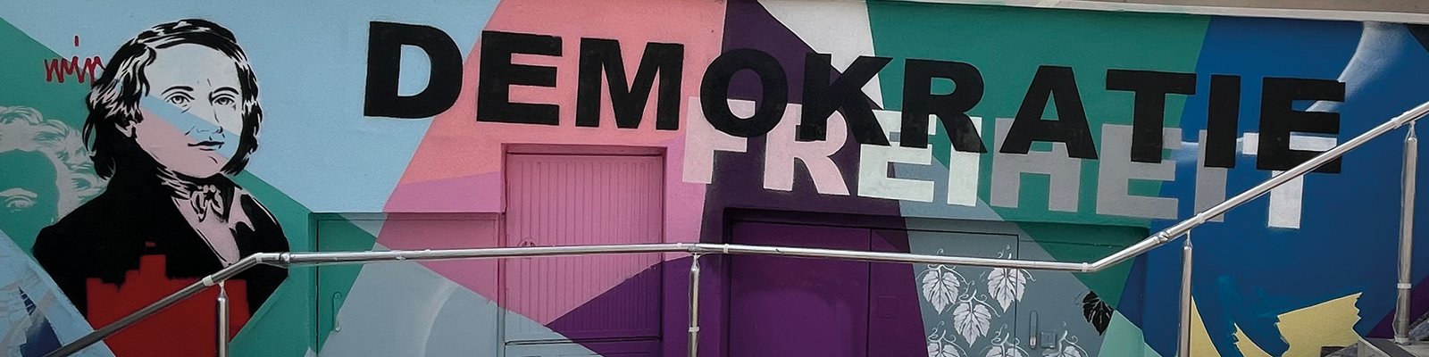 Zu sehen ist eine Graffitiwand. Viele bunte Farben, linke Seite eine schwarz-wei&szlig; Kontur eines Mannes. Quer dar&uuml;ber steht in gro&szlig;em Buchstaben: Demokratie Freiheit