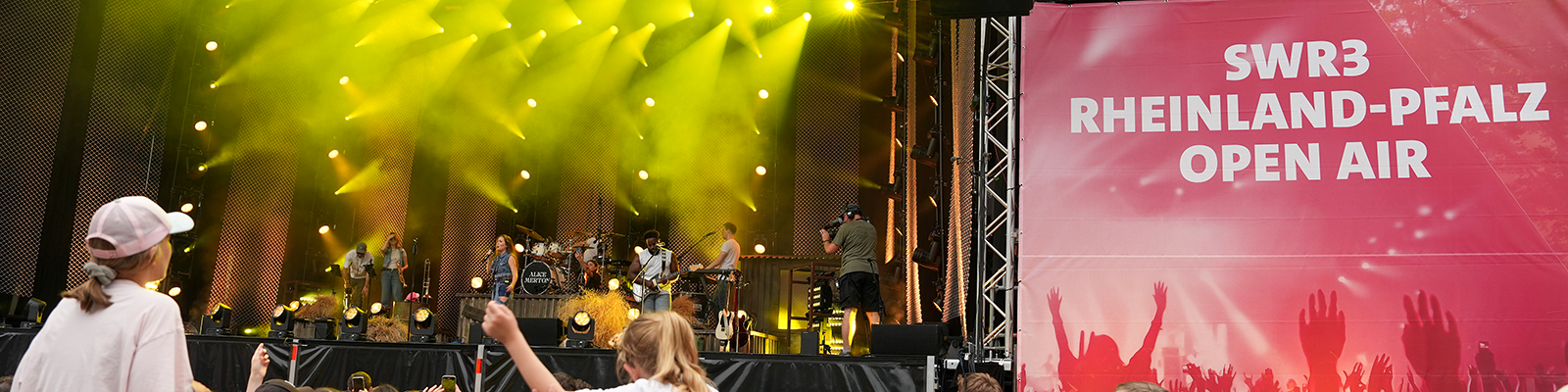Zu sehen ist eine Band auf einer großen Bühne. Davor sieht man viele Menschen.