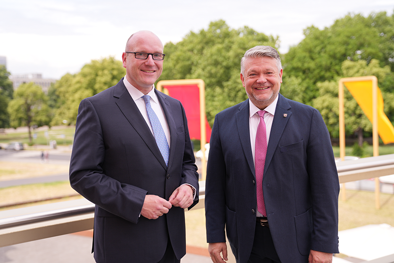 Zu sehen sind zwei Männer, im Hintergrund sieht man das Kunstobjekt der Fahnen in schwarz, rot, gold.