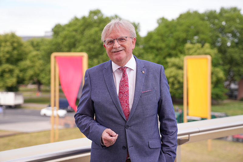 Zu sehen ist ein Mann, im Hintergrund sieht man das Kunstobjekt der Fahnen in schwarz, rot und gold.