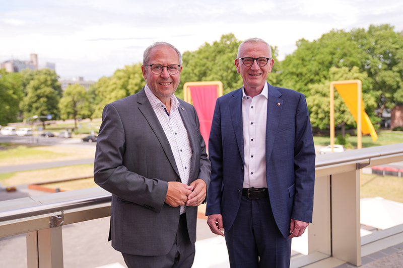 Zu sehen sind zwei Männer, im Hintergrund sieht man das Kunstobjekt der Fahnen in schwarz, rot, gold.